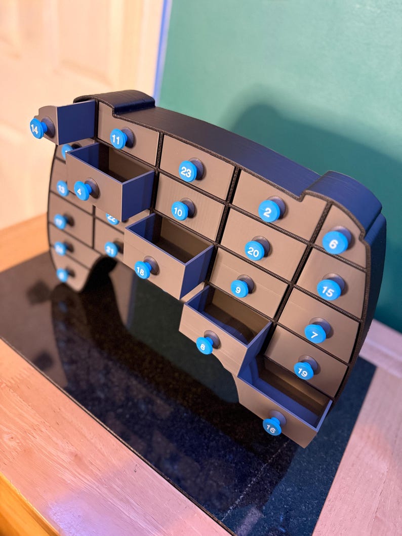 May include: A gray and blue advent calendar with 24 numbered drawers. Each drawer has a blue knob. The calendar is shaped like a rounded rectangle and sits on a dark surface.