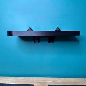 May include: A black floating shelf with two brackets, mounted on a teal wall. The shelf is made of wood and has a simple, modern design. The brackets are attached to the underside of the shelf and are designed to hold items. The shelf is approximately 61 cm long.