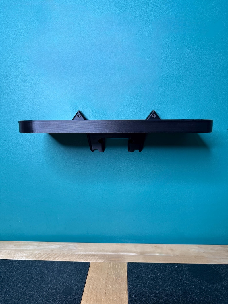 May include: A black floating shelf with two brackets, mounted on a teal wall. The shelf is made of wood and has a simple, modern design. The brackets are attached to the underside of the shelf and are designed to hold items. The shelf is approximately 61 cm long.