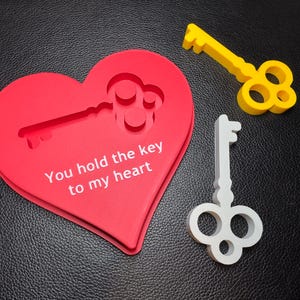 This is a heart-shaped red 3D-printed decoration featuring a key-shaped cutout and the engraved text ‘You hold the key to my heart.’ Alongside it are two keys, one gold and one silver, designed to fit the cutout in the heart.