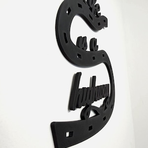 May include: Black wall art in the shape of a horseshoe with the words "we are a highway" in a cursive font. The art piece is made of a textured material and is mounted on a white wall.