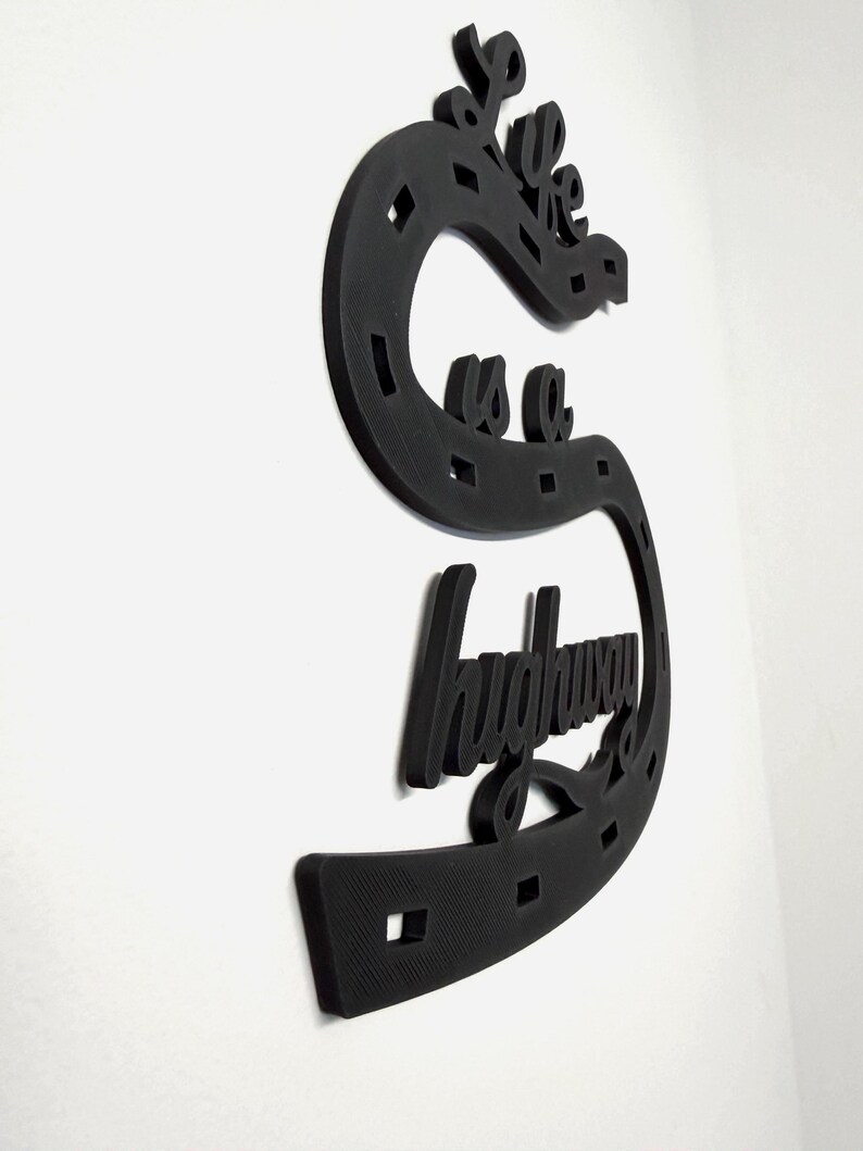 May include: Black wall art in the shape of a horseshoe with the words "we are a highway" in a cursive font. The art piece is made of a textured material and is mounted on a white wall.