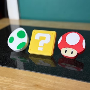 May include: Three colorful, 3D-printed Super Mario Bros. themed items: a white egg with green spots, a yellow question mark block, and a red mushroom with white spots. The items are arranged on a dark surface.