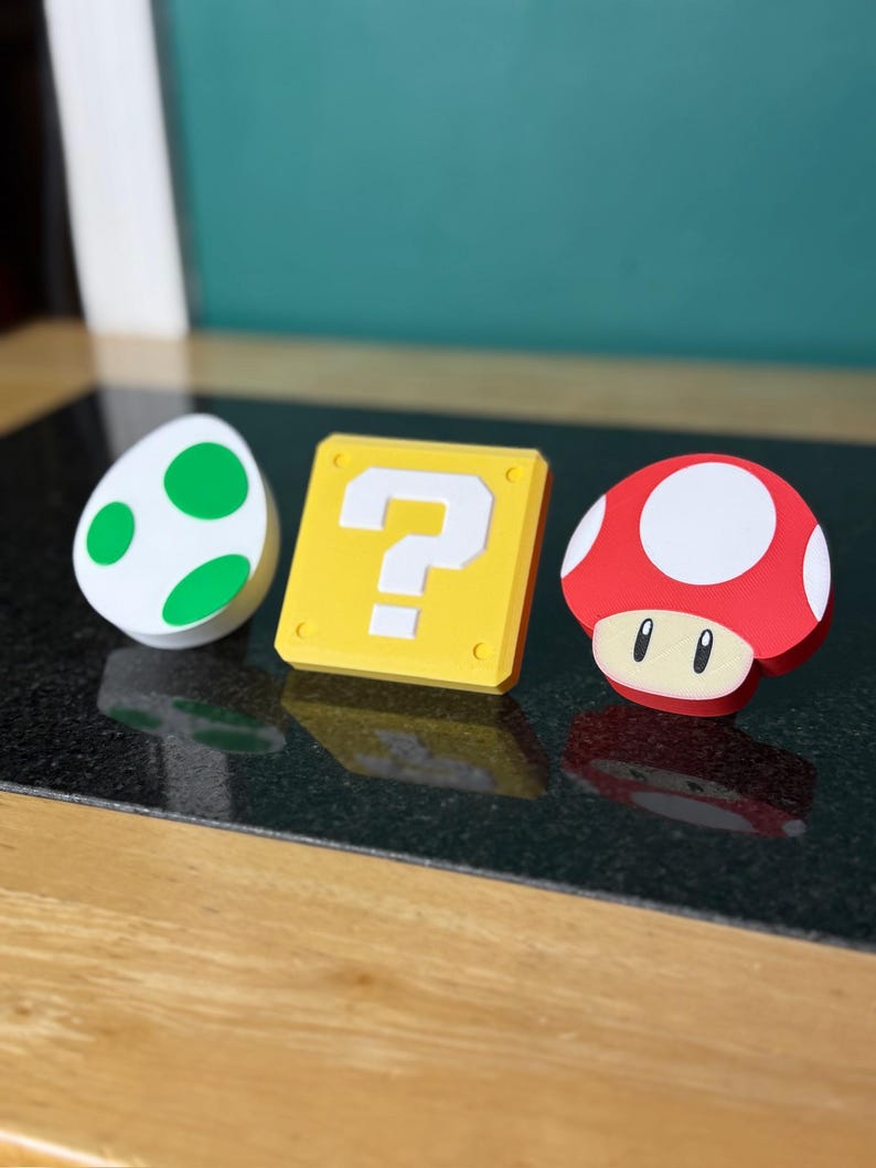 May include: Three colorful, 3D-printed Super Mario Bros. themed items: a white egg with green spots, a yellow question mark block, and a red mushroom with white spots. The items are arranged on a dark surface.