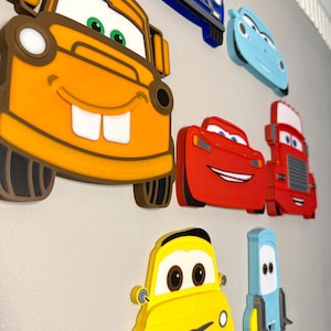 May include: A collection of layered wall-mounted decorations featuring cartoon car characters. The set includes a large orange tow truck, a red sports car, a red truck, a yellow car, a light blue car, a dark blue car, and a small light blue vehicle.