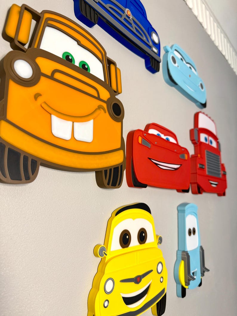 May include: A collection of layered wall-mounted decorations featuring cartoon car characters. The set includes a large orange tow truck, a red sports car, a red truck, a yellow car, a light blue car, a dark blue car, and a small light blue vehicle.