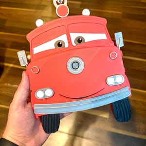 K&ouml;nnte beinhalten: Ein rotes, cartoonf&ouml;rmiges Autozubeh&ouml;r mit einem l&auml;chelnden Gesicht, Scheinwerfern und Seitenspiegeln. Das Zubeh&ouml;r hat schwarze Reifen und eine wei&szlig;e und silberne Zierleiste. Das Auto hat ein kleines wei&szlig;es und rotes Emblem oben.