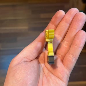 Puede incluir: Un peque&ntilde;o componente de pl&aacute;stico amarillo y negro. El objeto tiene una base negra rectangular y un cuerpo amarillo con una serie de crestas verticales. El objeto se sostiene en una mano.