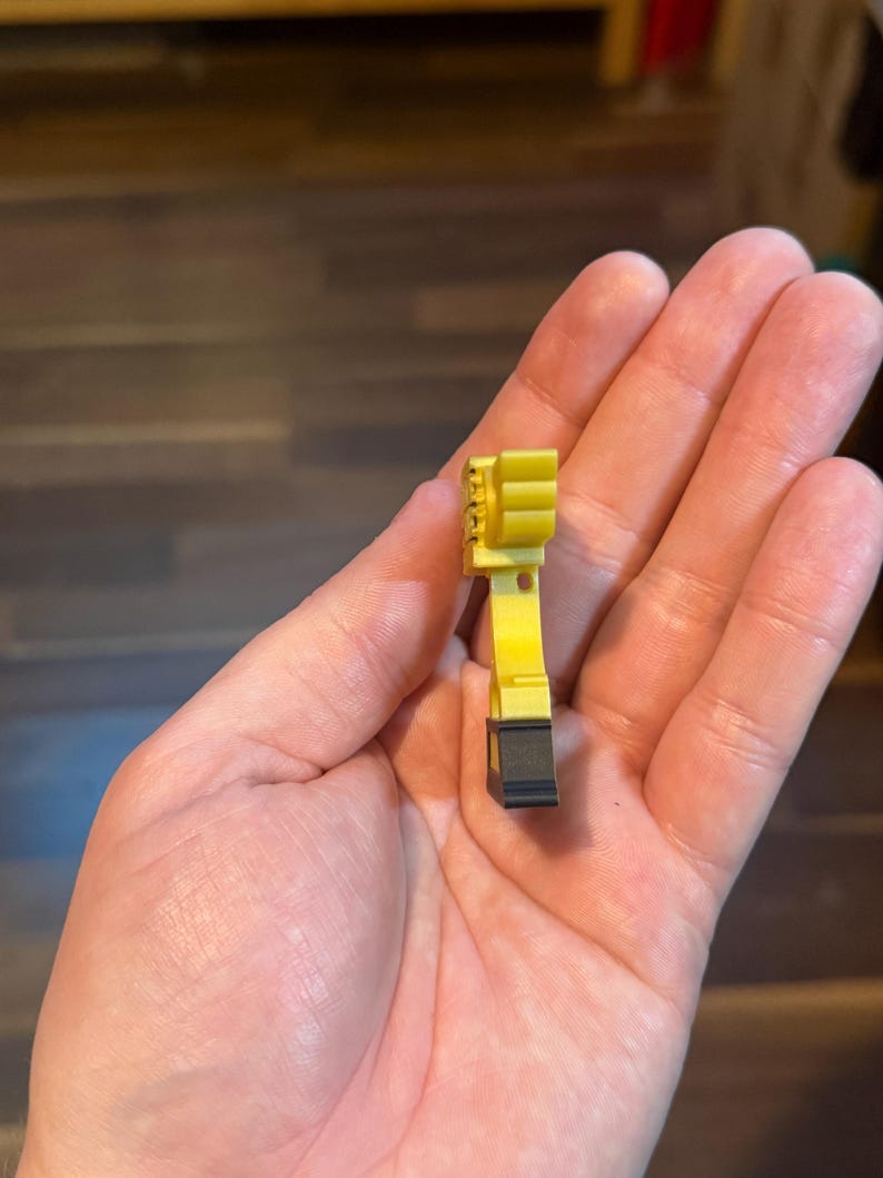 Puede incluir: Un peque&ntilde;o componente de pl&aacute;stico amarillo y negro. El objeto tiene una base negra rectangular y un cuerpo amarillo con una serie de crestas verticales. El objeto se sostiene en una mano.