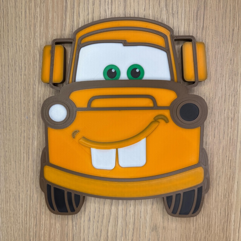 May include: Orange and brown cartoon car character with a smiling face, white teeth, and green eyes. The car has a detailed design with headlights, a windshield, and side mirrors. The character is a popular children's toy.
