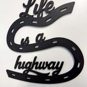May include: A black wall decoration shaped like a winding road with dashed white-like markings. The text "Life is a highway" is integrated into the design in a cursive script font. The object is flat and features a continuous, curved path.