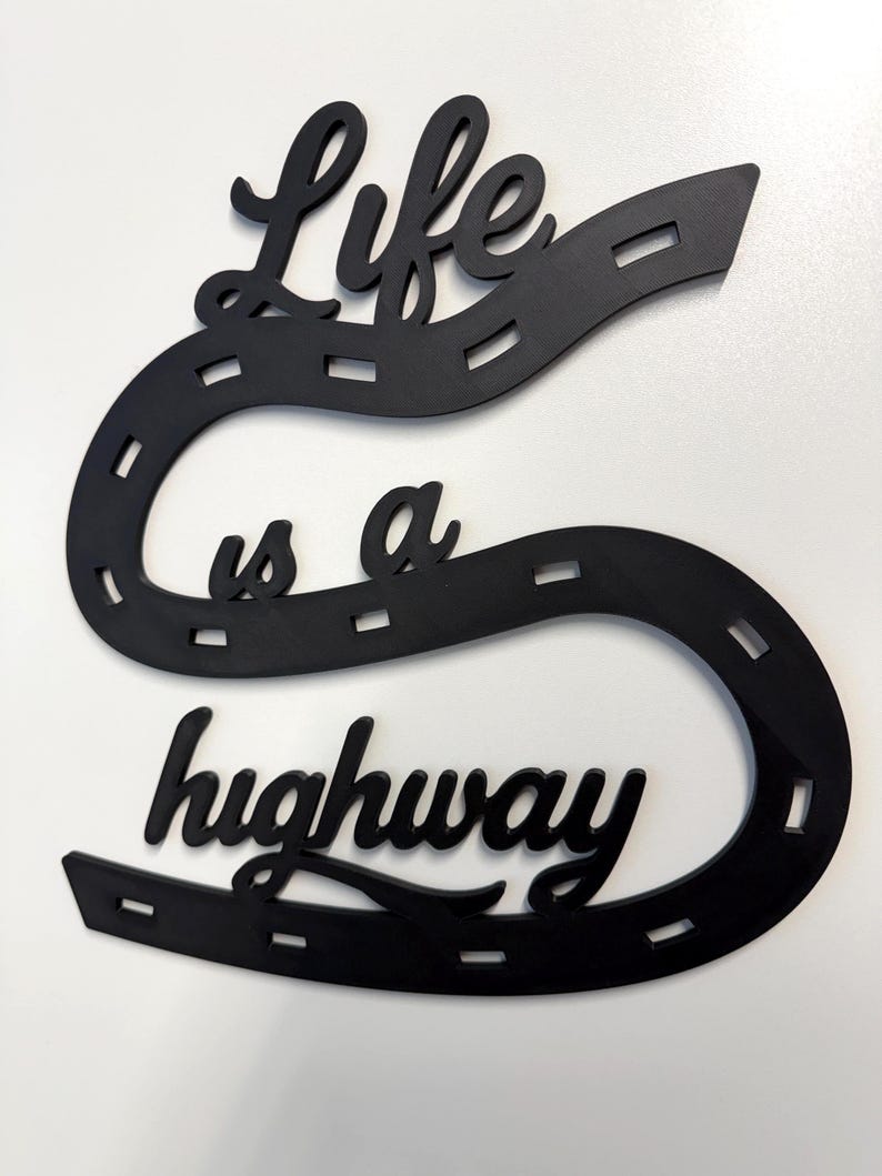 May include: A black wall decoration shaped like a winding road with dashed white-like markings. The text "Life is a highway" is integrated into the design in a cursive script font. The object is flat and features a continuous, curved path.