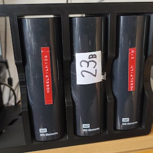 K&ouml;nnte beinhalten: Ein schwarzer Kunststoff-Aufbewahrungsst&auml;nder mit drei vertikalen WD Elements externen Festplatten. Die beiden &auml;u&szlig;eren Laufwerke haben rote Etiketten mit dem Text HOSELFILM, das mittlere Laufwerk ein wei&szlig;es Etikett mit der Aufschrift 23B.