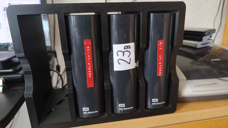 K&ouml;nnte beinhalten: Ein schwarzer Kunststoff-Aufbewahrungsst&auml;nder mit drei vertikalen WD Elements externen Festplatten. Die beiden &auml;u&szlig;eren Laufwerke haben rote Etiketten mit dem Text HOSELFILM, das mittlere Laufwerk ein wei&szlig;es Etikett mit der Aufschrift 23B.