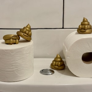 Gold glittery 3D-printed poop emojis displayed on toilet rolls atop a toilet cistern. A humorous, eco-friendly decoration made from recycled filament waste, adding a playful touch to any bathroom.