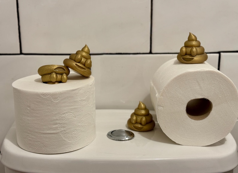 Gold glittery 3D-printed poop emojis displayed on toilet rolls atop a toilet cistern. A humorous, eco-friendly decoration made from recycled filament waste, adding a playful touch to any bathroom.