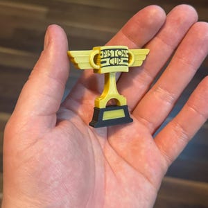 Puede incluir: Un trofeo de la Copa Pist&oacute;n amarillo y negro con el texto "Piston Cup" en la parte delantera. El trofeo tiene alas a cada lado y se sostiene en una mano. Es una r&eacute;plica en miniatura.