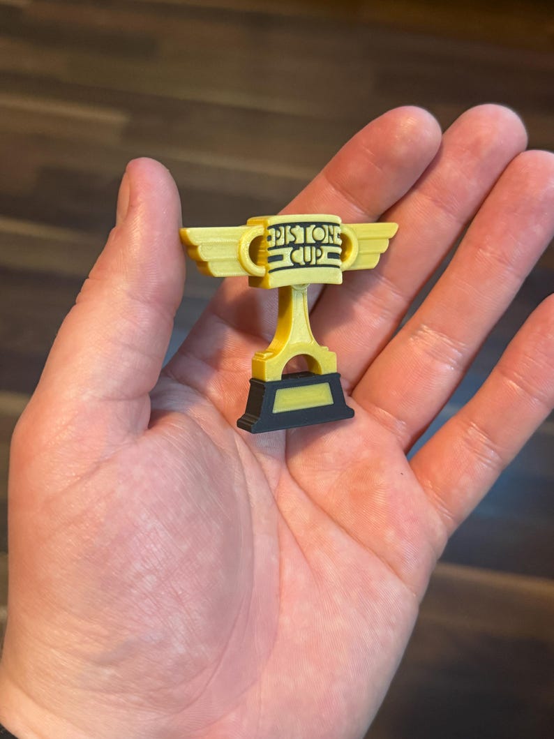 Puede incluir: Un trofeo de la Copa Pist&oacute;n amarillo y negro con el texto "Piston Cup" en la parte delantera. El trofeo tiene alas a cada lado y se sostiene en una mano. Es una r&eacute;plica en miniatura.