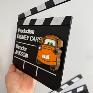 May include: A black and white film clapperboard with the text "Production DISNEY CARS" and a cartoon car character. The clapperboard is held up, with a second clapperboard visible in the background. The clapperboard is a movie-themed item.