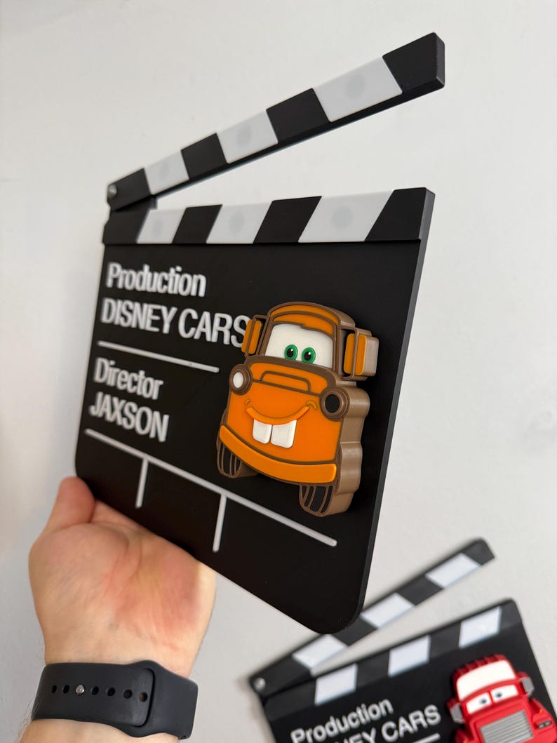 May include: A black and white film clapperboard with the text "Production DISNEY CARS" and a cartoon car character. The clapperboard is held up, with a second clapperboard visible in the background. The clapperboard is a movie-themed item.