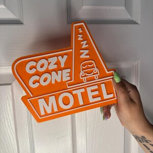 以下が含まれることがあります： オレンジと白の看板に「Cozy Cone Motel」の文字と、ZZZが書かれた漫画の車が描かれている