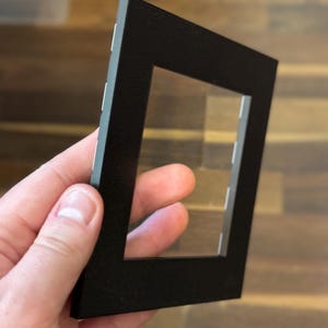 May include: A black picture frame with a rectangular opening and a clear plastic insert. The frame is held at an angle, showcasing its clean lines and matte finish. The frame is designed to hold a photograph or artwork.