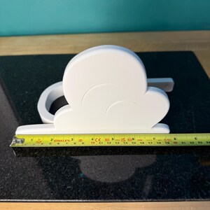 May include: A white cloud-shaped decorative object, approximately 9 inches long, with a curved design and a flat base. The object is placed on a dark, speckled surface, with a yellow measuring tape alongside it.
