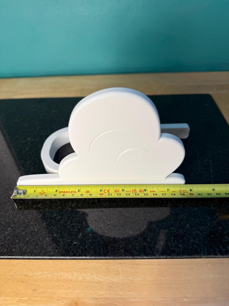 May include: A white cloud-shaped decorative object, approximately 9 inches long, with a curved design and a flat base. The object is placed on a dark, speckled surface, with a yellow measuring tape alongside it.