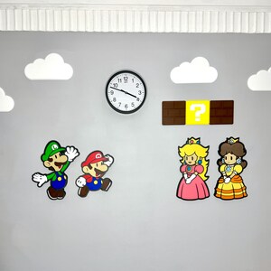 Op de afbeelding: Een wanddecoratie met personages uit een populair videospel. De sc&egrave;ne omvat een zwarte klok, stripfiguren, een vraagtekenblok en witte wolkenuitsparingen tegen een grijze muur. De personages zijn Mario, Luigi, Princess Peach en Daisy.