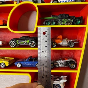 May include: A red and yellow toy car display shelf filled with various toy cars. The shelf has a yellow frame and red interior. The toy cars are of different colors and designs. A ruler shows the shelf is about 4 inches tall.