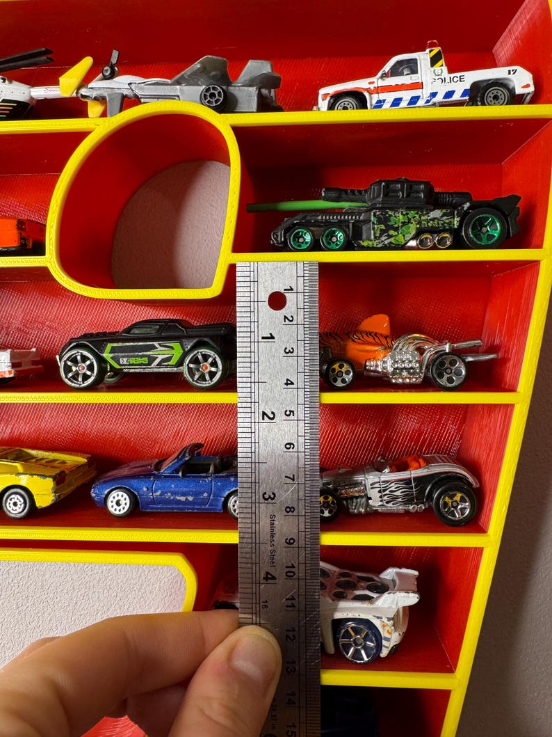 May include: A red and yellow toy car display shelf filled with various toy cars. The shelf has a yellow frame and red interior. The toy cars are of different colors and designs. A ruler shows the shelf is about 4 inches tall.