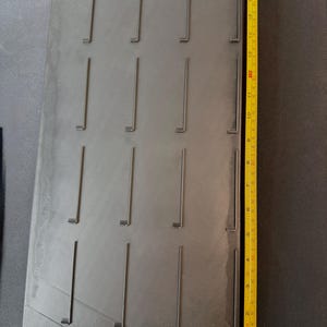 May include: A dark gray rectangular display board with multiple vertical slots. A yellow measuring tape shows the board is approximately 16 inches long. The board has a textured surface and a checkered pattern at the bottom.
