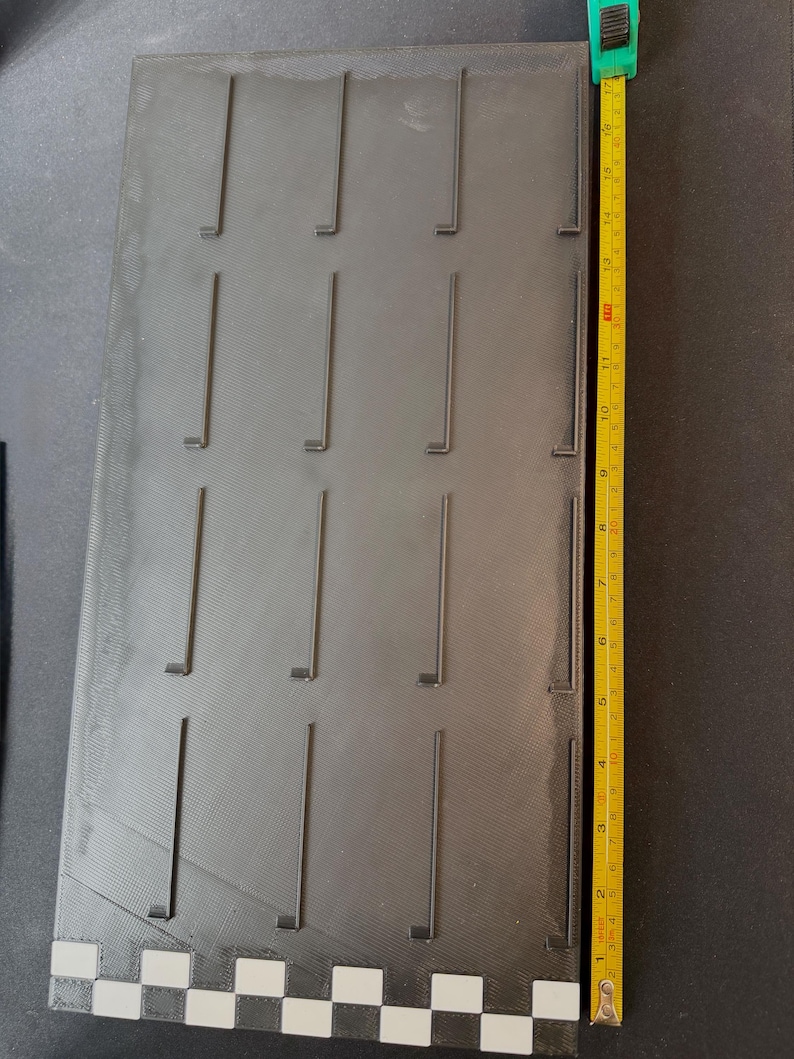 May include: A dark gray rectangular display board with multiple vertical slots. A yellow measuring tape shows the board is approximately 16 inches long. The board has a textured surface and a checkered pattern at the bottom.