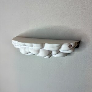 May include: A white cloud-shaped shelf mounted on a wall. The shelf has a rounded, textured design, resembling fluffy clouds. The shelf is made of a solid, opaque material and is designed to hold small items. The shelf is approximately 12 inches long.
