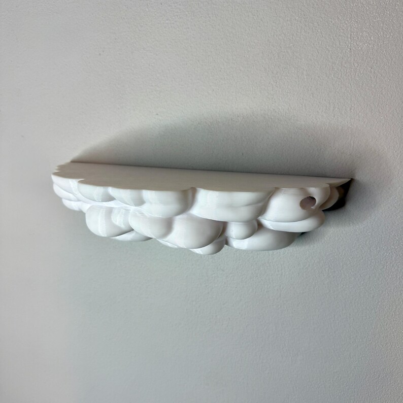 May include: A white cloud-shaped shelf mounted on a wall. The shelf has a rounded, textured design, resembling fluffy clouds. The shelf is made of a solid, opaque material and is designed to hold small items. The shelf is approximately 12 inches long.