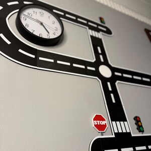 May include: A black and white road-themed wall decal with a clock, stop sign, and traffic lights. The clock has a black frame and white face. The road design includes a roundabout and crosswalk.