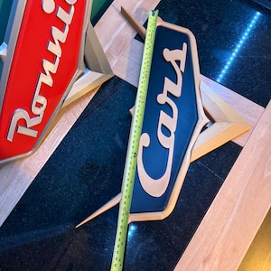 May include: Two vintage-style signs, one red with "Ronnie" and the other blue with "Cars" in a stylized font. The signs are approximately 20 inches long. The signs have a gold-colored base and are likely for a car-related business.