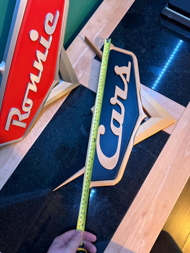 May include: Two vintage-style signs, one red with "Ronnie" and the other blue with "Cars" in a stylized font. The signs are approximately 20 inches long. The signs have a gold-colored base and are likely for a car-related business.