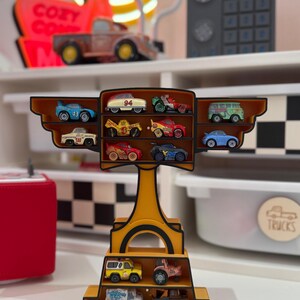 Puede incluir: Una estanter&iacute;a amarilla llena de coches de juguete en miniatura. La estanter&iacute;a tiene m&uacute;ltiples niveles y compartimentos, mostrando varios veh&iacute;culos coloridos. Los coches incluyen coches de carreras, camiones y furgonetas. La estanter&iacute;a est&aacute; decorada con un borde negro y marr&oacute;n.