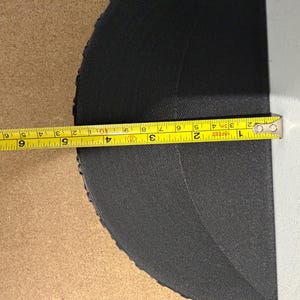 Może przedstawiać: Czarny, p&oacute;łokrągły przedmiot jest mierzony ż&oacute;łtą miarką. Miarka pokazuje długość około 10 cm. Przedmiot wydaje się być wykonany z teksturowanego materiału.