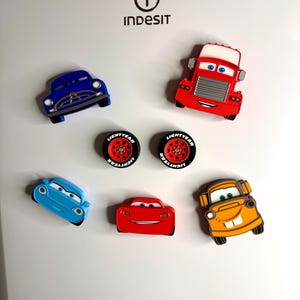 May include: A collection of colorful car-shaped magnets. The magnets include a blue car, a red truck, two tires, a light blue car, a red car, and an orange car. The magnets are designed with cartoon-like features.