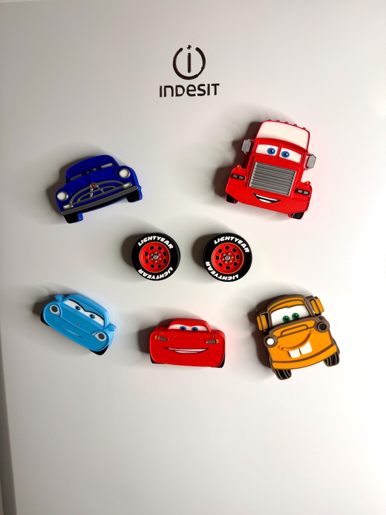 May include: A collection of colorful car-shaped magnets. The magnets include a blue car, a red truck, two tires, a light blue car, a red car, and an orange car. The magnets are designed with cartoon-like features.