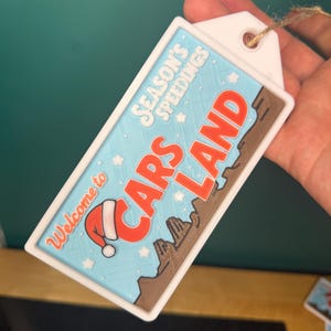 K&ouml;nnte beinhalten: Ein rechteckiges wei&szlig;es Etikett mit blauem Hintergrund mit dem Text "Welcome to Cars Land" und "Season's Speedings". Das Etikett hat einen roten Weihnachtsmannhut und ein braunes W&uuml;stenlandschaftsdesign. Ein Bindfaden ist am Etikett befestigt.