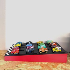 May include: A display stand with a red base and a black top featuring a checkered flag design. The stand holds a collection of colorful toy cars, including red, yellow, blue, and green vehicles, arranged in rows.