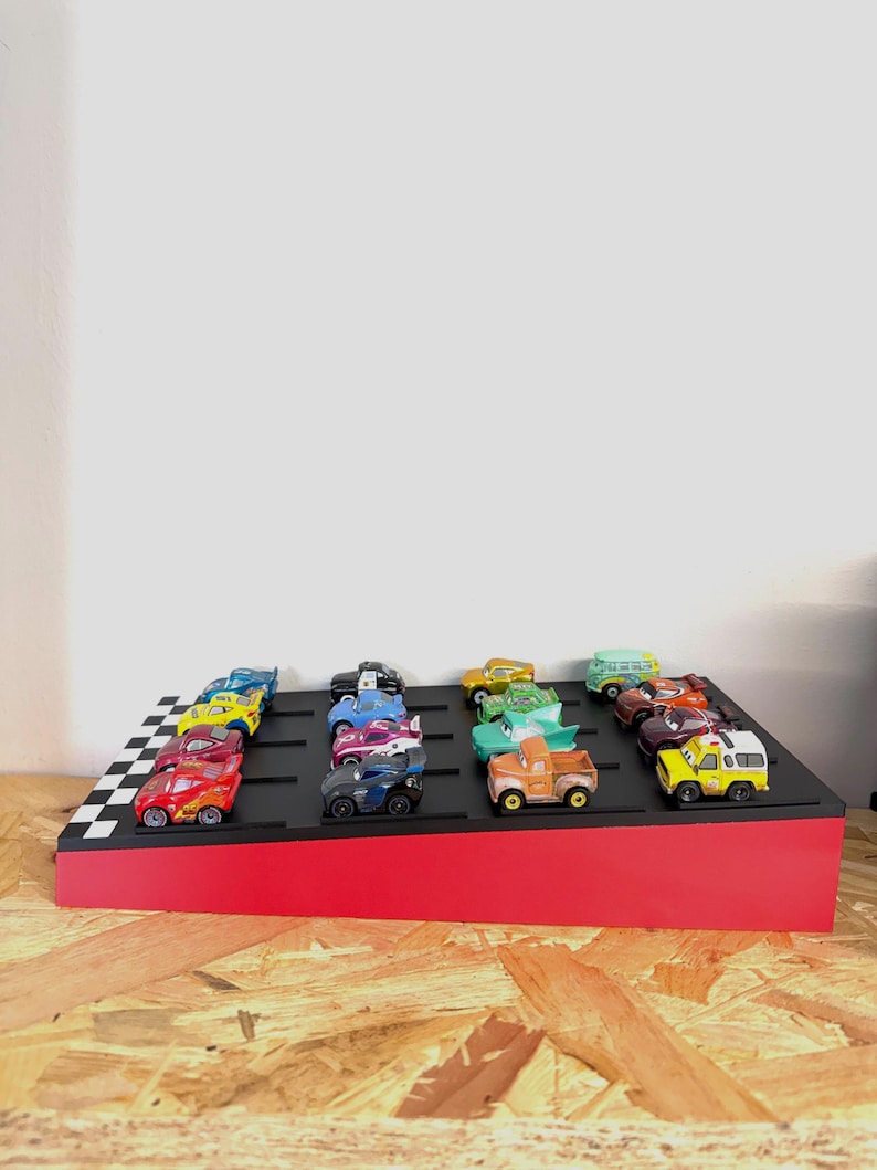 May include: A display stand with a red base and a black top featuring a checkered flag design. The stand holds a collection of colorful toy cars, including red, yellow, blue, and green vehicles, arranged in rows.