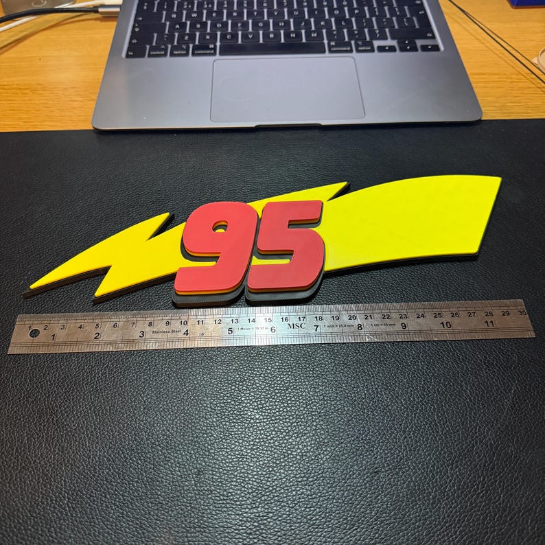 May include: A yellow lightning bolt with the number 95 in red, with a black outline. The number is approximately 10 inches long. The design is on a black surface, with a laptop and ruler in the background.