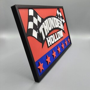 May include: A rectangular sign with a black frame, featuring the words "THUNDER HOLLOW" in white, with checkered flags and red stars on a red and blue background. The sign is angled.