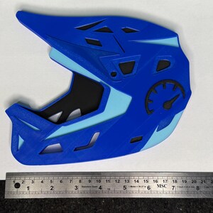 May include: A blue and light blue 3D-printed helmet with a speedometer graphic. The helmet has a visor and multiple vents. A metal ruler is at the bottom of the image.