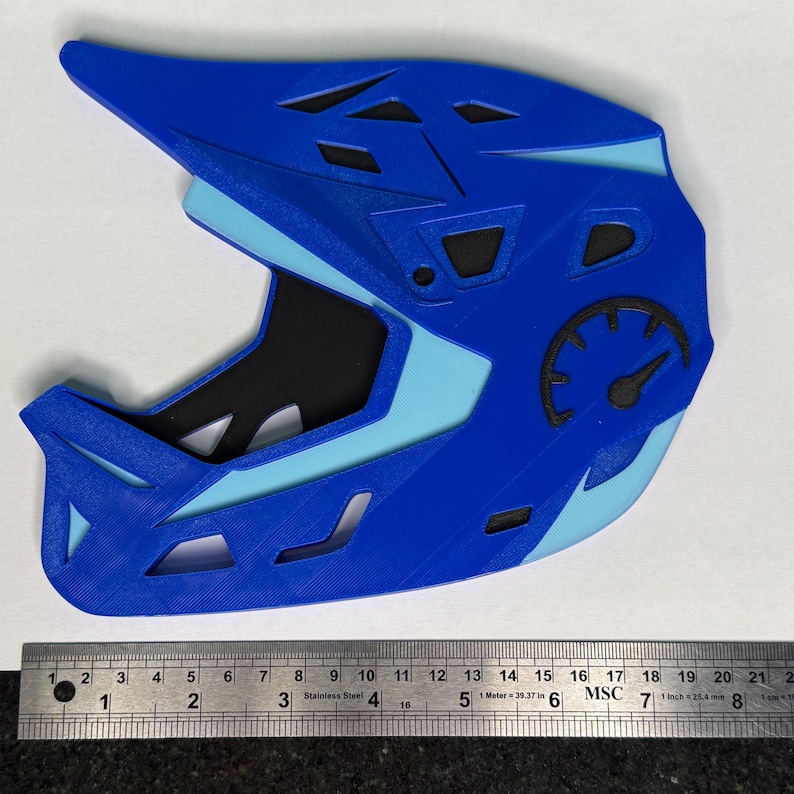 May include: A blue and light blue 3D-printed helmet with a speedometer graphic. The helmet has a visor and multiple vents. A metal ruler is at the bottom of the image.