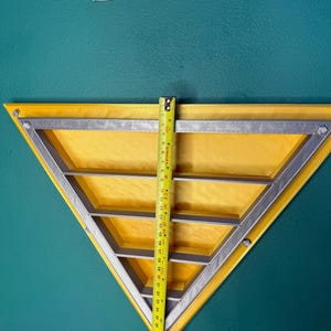 May include: A yellow triangular shelf with a silver metal frame and three interior shelves. A yellow tape measure shows the shelf is approximately 30 inches tall. The shelf is mounted on a teal wall.